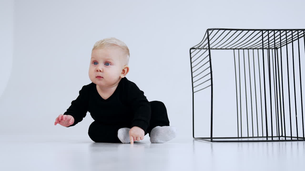 Grey-eyed blond baby in black suit sits on the floor near the metal cake. Kid stands on all four and crawls by the floor away. White backdrop.