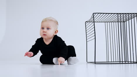 grey-eyed blond baby in black suit sits on the floor near the metal cake. kid stands on all four and crawls by the floor away. white backdrop.