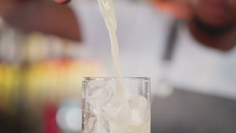 barman pours exotic cocktail into glass in bar closeup. african american servant fills goblet with cocktail in nightclub. delicious beverage with ice cubes