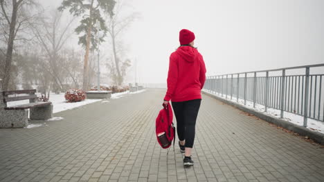 vista trasera de un atleta con chaqueta roja de invierno y calentador de cabeza, sosteniendo una bolsa, caminando a través de un paisaje nevado cubierto de niebla con árboles y bancos cubiertos de nieve