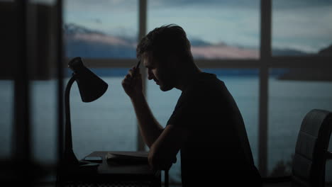 a thoughtful man sitting at a table with a desk lamp writes with a pen. dark time of the day twilight on the background of the window a sad man thinks and writes