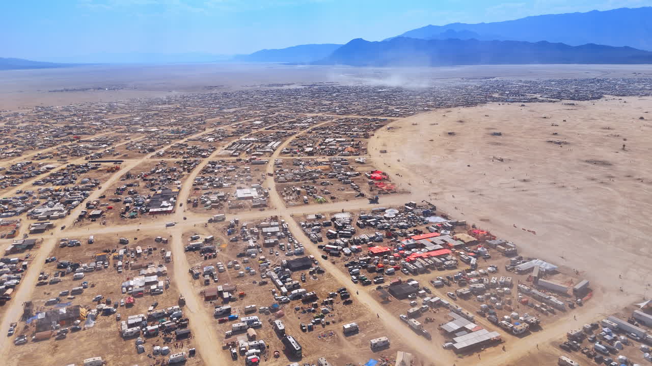 Wide panorama of temporary desert city and mountain range. Extreme wide aerial view of an expansive temporary settlement in a dry valley with distant mountains and blue sky