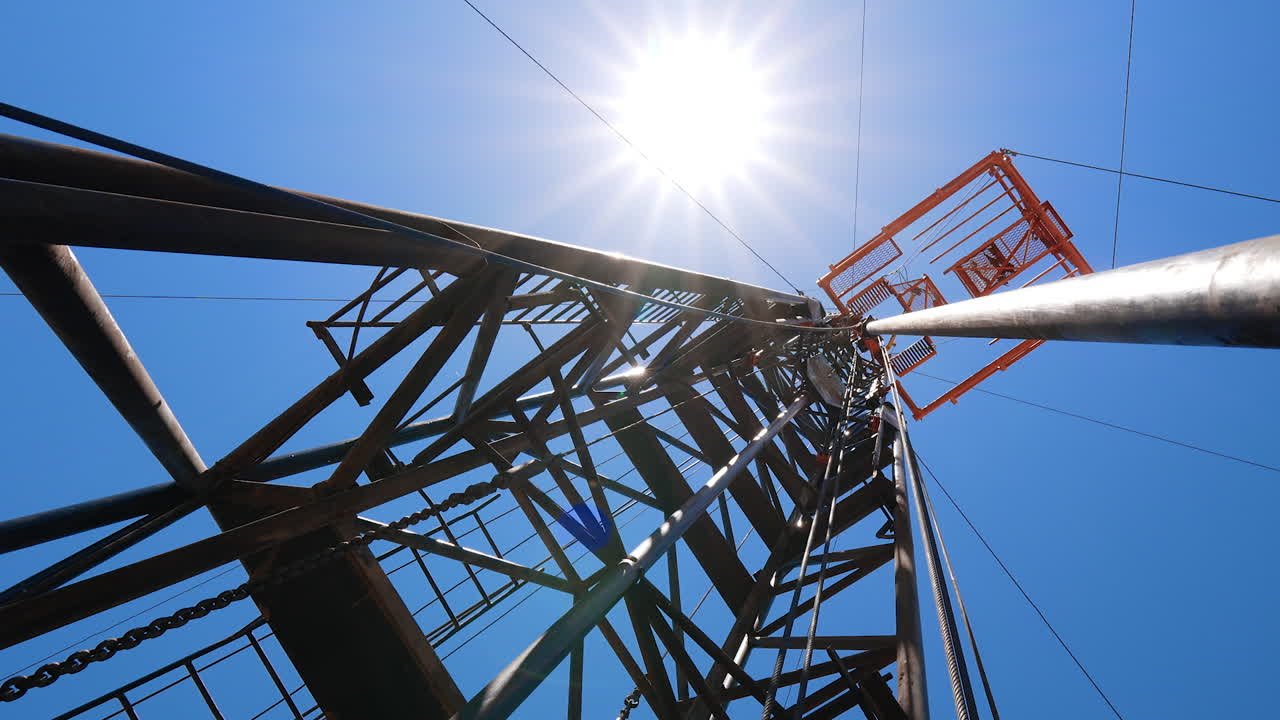Metal wires hang from the oil derrick. Low angle view at the tower with the boer for producing natural resources.