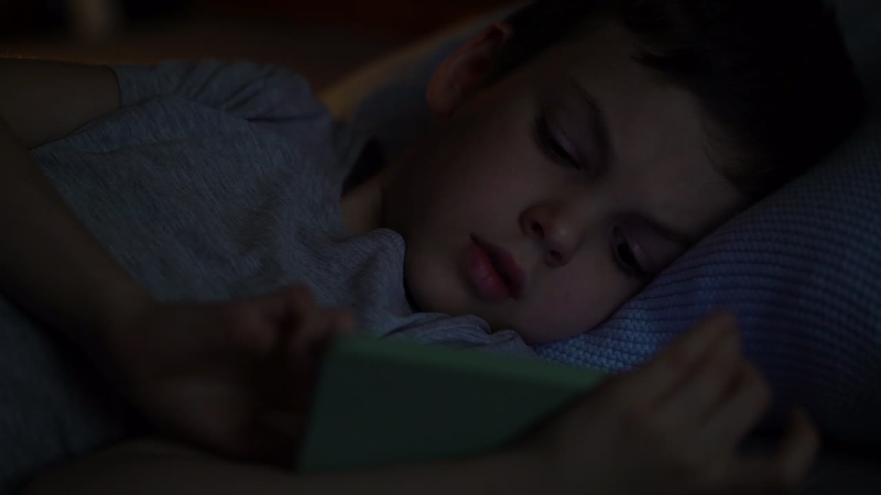 Bored preschooler lying on his bed in the dark and used app on his phone.