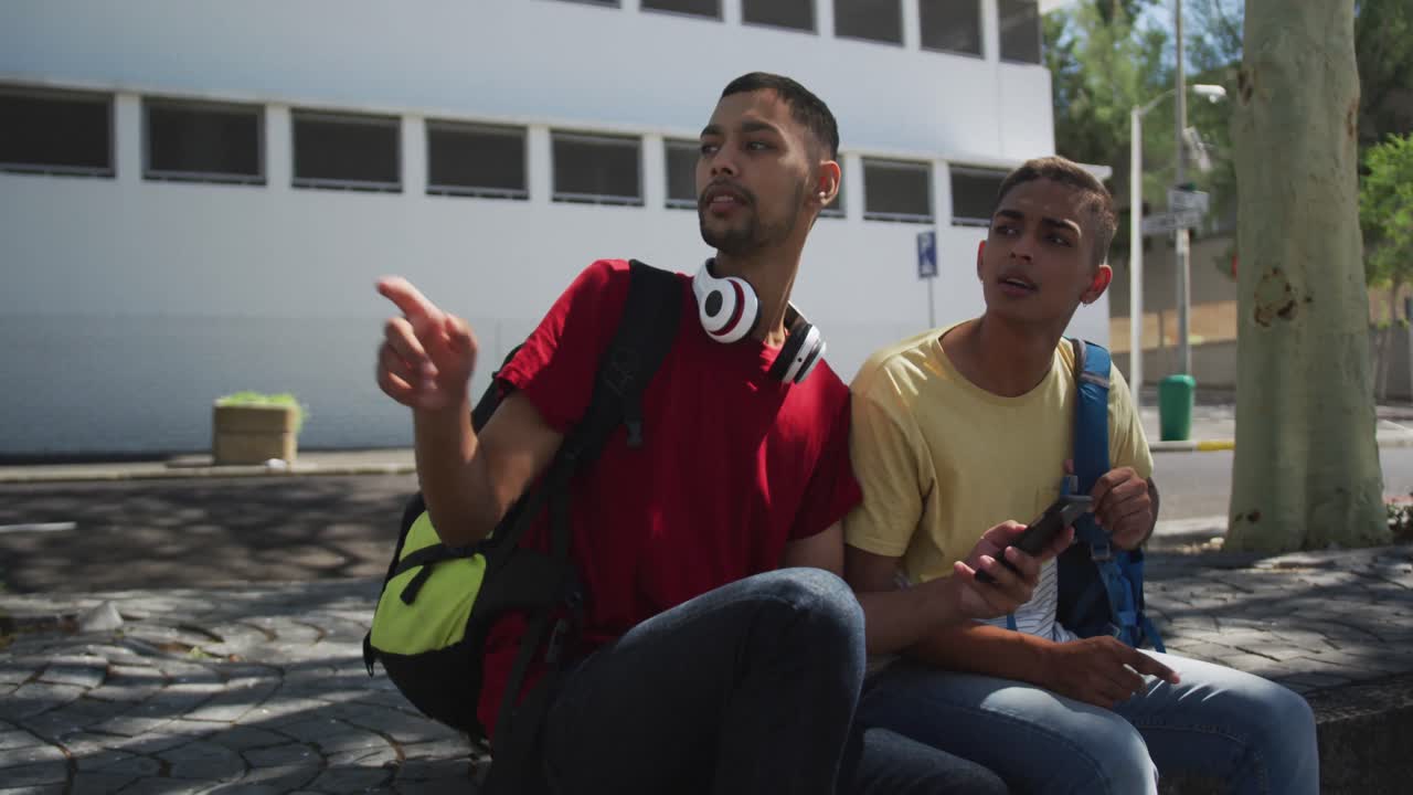 dos amigos de raza mixta sentados y usando un teléfono inteligente en la calle