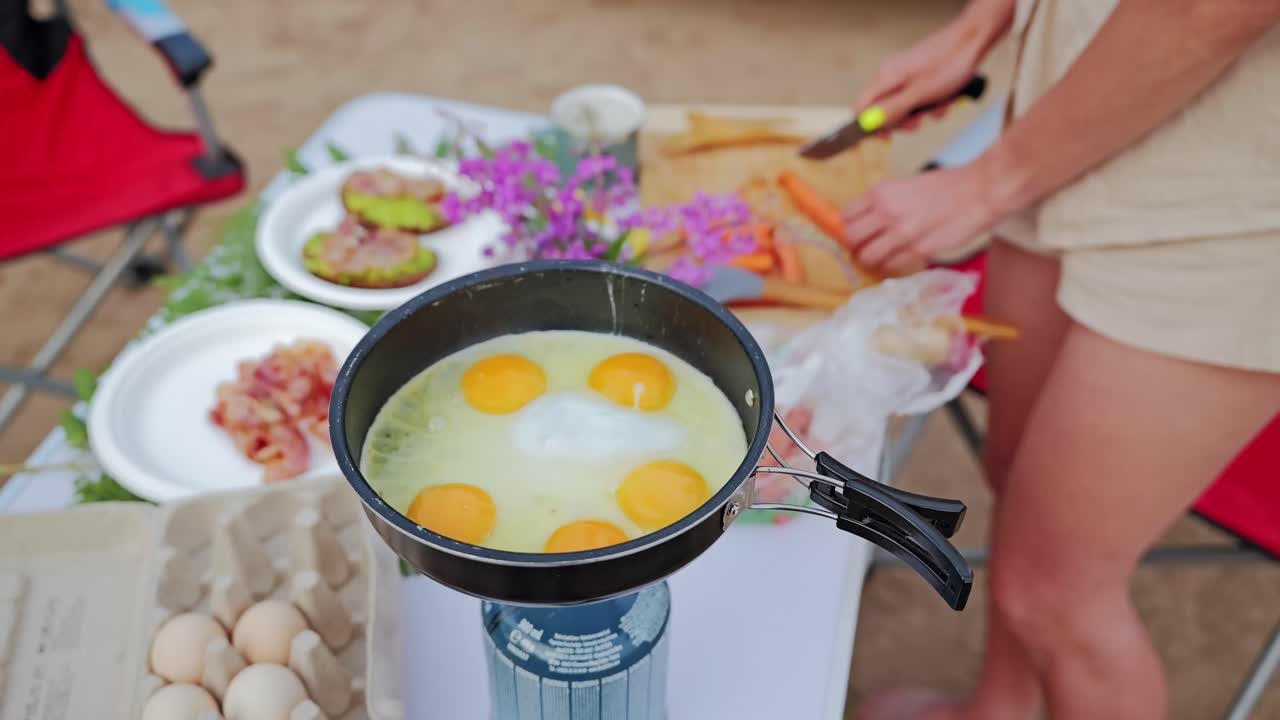 Outdoor cooking scene with eggs in pan and woman prepping food in background