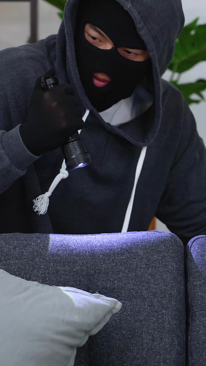 A criminal in a balaclava uses a flashlight to search a dark living room interior