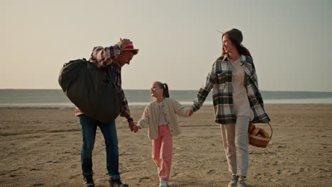 a happy brunette man with gray hair in a checkered shirt carries a large green bag and walks with his wife, a brunette girl in a green checkered shirt, and with his little daughter, a blonde girl in a white jacket, near the seashore during his hike and picnic