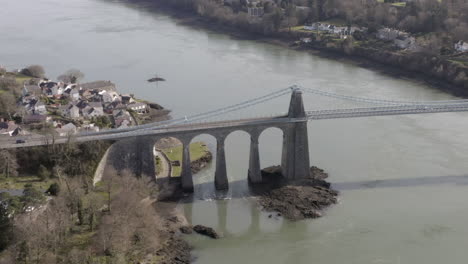 Aerial-view-of-Menai-Suspension-Bridge,-flying-right-to-left-around-the-bridge-while-zooming-out,-Anglesey,-North-Wales,-UK