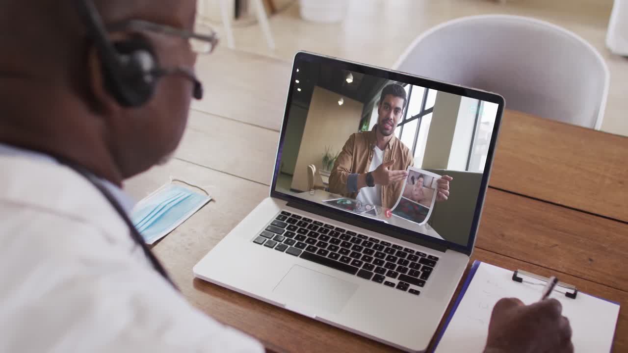 Afroamerikansk mandlig læge med headset på, der tager noter under et videoopkald på sin bærbare computer