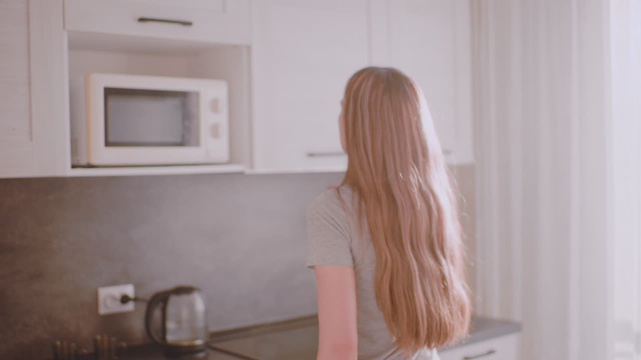Una giovane donna entra in cucina, vista dal corridoio, e raggiunge il pensile, prende il contenitore del caffè, lo appoggia sul piano di lavoro e poi sposta la tazza in avanti con il bollitore in vista