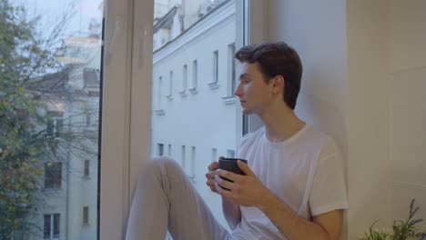 sliding portrait of pensive man drinking coffee alone by window on autumn background. side view of sad boy sitting at windowsill indoors holding tea cup slow motion. depression loneliness isolation