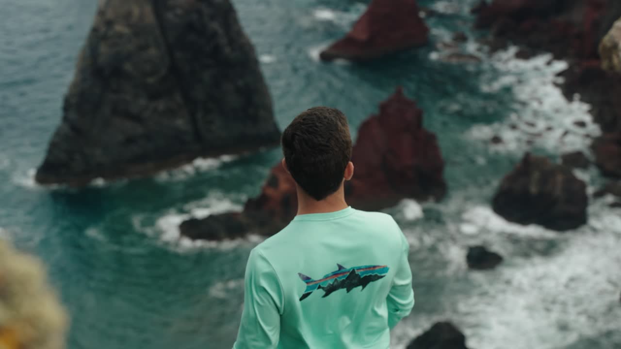 A man in a turquoise shirt stands on a cliff edge, gazing at the jagged volcanic rocks and turquoise waves below.