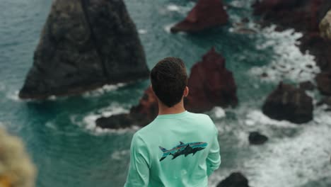 a man in a turquoise shirt stands on a cliff edge, gazing at the jagged volcanic rocks and turquoise waves below.