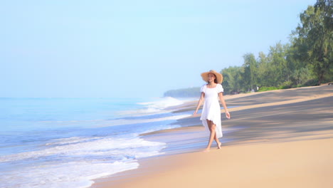 Mientras-Las-Olas-Avanzan-A-Lo-Largo-De-La-Playa,-Una-Mujer-Joven-Con-Un-Vestido-De-Verano-Y-Un-Sombrero-De-Paja-Pasea-Por-El-Borde-De-Las-Olas