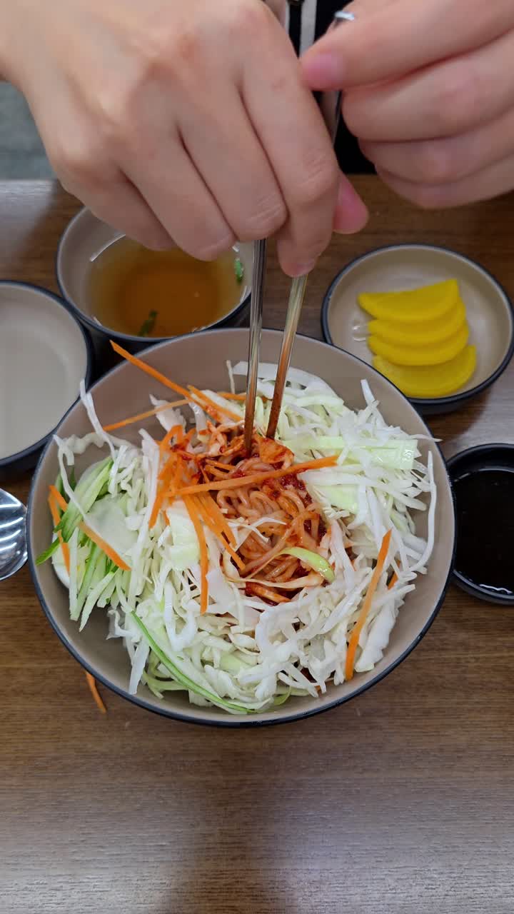 A person uses chopsticks to mix a bowl of Korean Jjolmyeon, combining chewy noodles with fresh shredded cabbage, vegetables, and a spicy gochujang sauce for a traditional and delicious meal