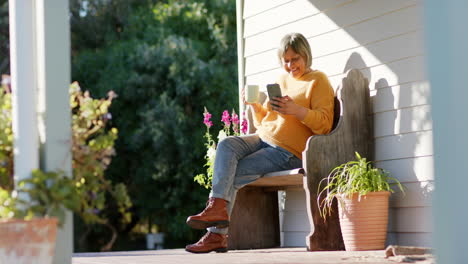 mujer biracial senior feliz usando teléfono inteligente y bebiendo café en la terraza, cámara lenta, espacio de copia