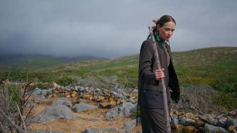 travel girl trekking hill with stick on cloudy day. serene grassy mountains view