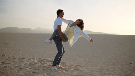 Perfect-scene-of-young-couple-in-empty-desert.-man-holding-her-woman-on-arms-and-turning-her-around,-have-fun-emotionally,-laugh,smile-on-sunset-at-desert-crazy-in-love,-emotions-and-relationship.-Slow-motion