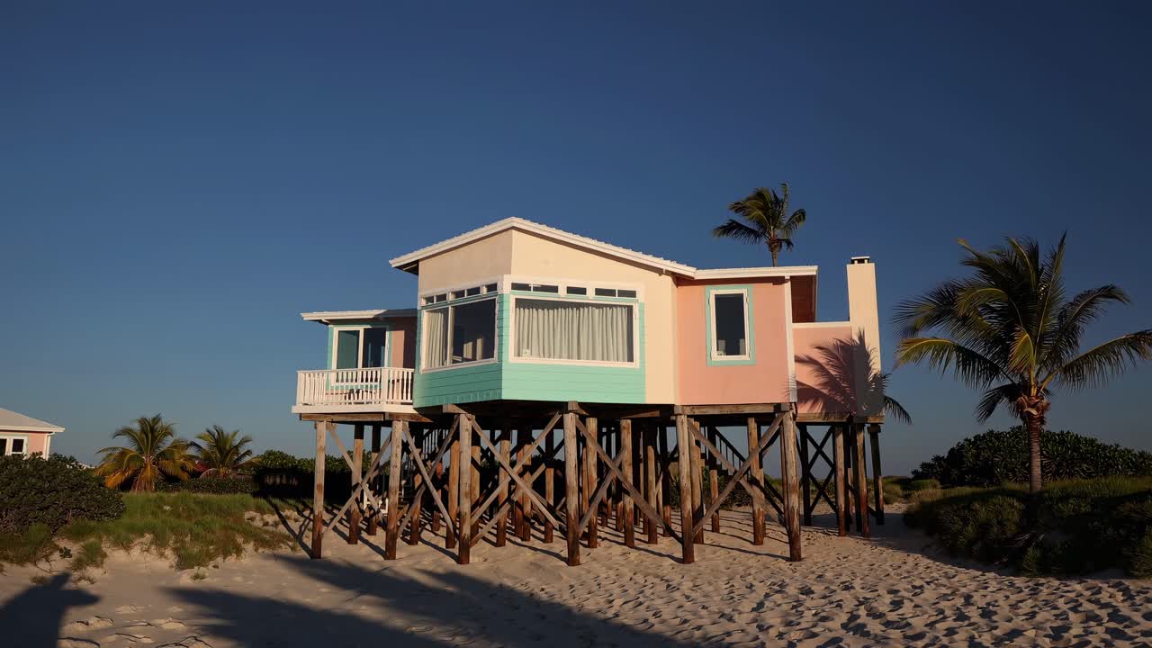 Pastel colored beach house elevated on wooden stilts, overlooking a pristine sandy beach with palm trees swaying gently in the tropical breeze, creating a serene and idyllic vacation setting