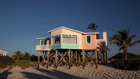 pastel colored beach house elevated on wooden stilts, overlooking a pristine sandy beach with palm trees swaying gently in the tropical breeze, creating a serene and idyllic vacation setting