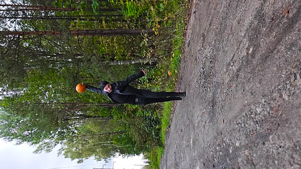 검은 옷을 입은 남자가 숲을 배경으로 한 시골 도로에서 하로윈 호박을 땅에 조각으로 던지는 수직 느린 동작 비디오