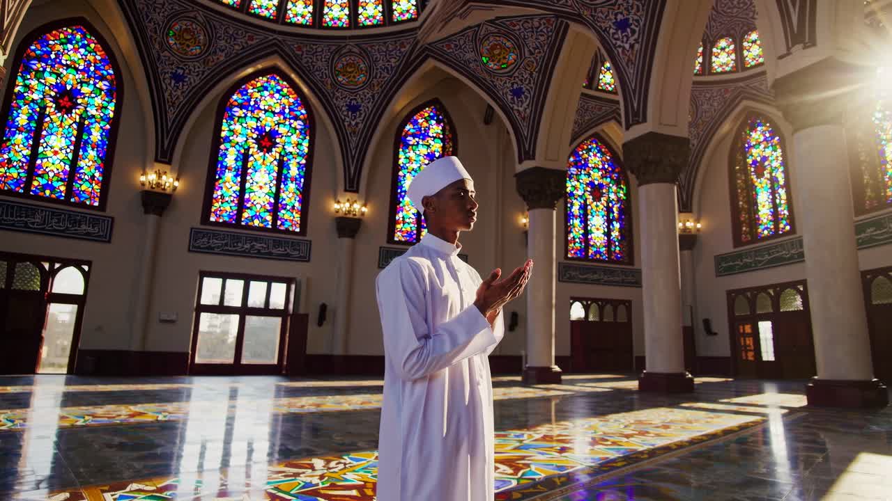 A man in traditional attire prays in a sunlit mosque with stained glass windows