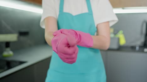 Close-up-of-a-confident-cleaning-lady-girl-in-a-blue-apron-and-white-T-shirt-putting-on-pink-rubber-gloves-for-cleaning-in-the-kitchen