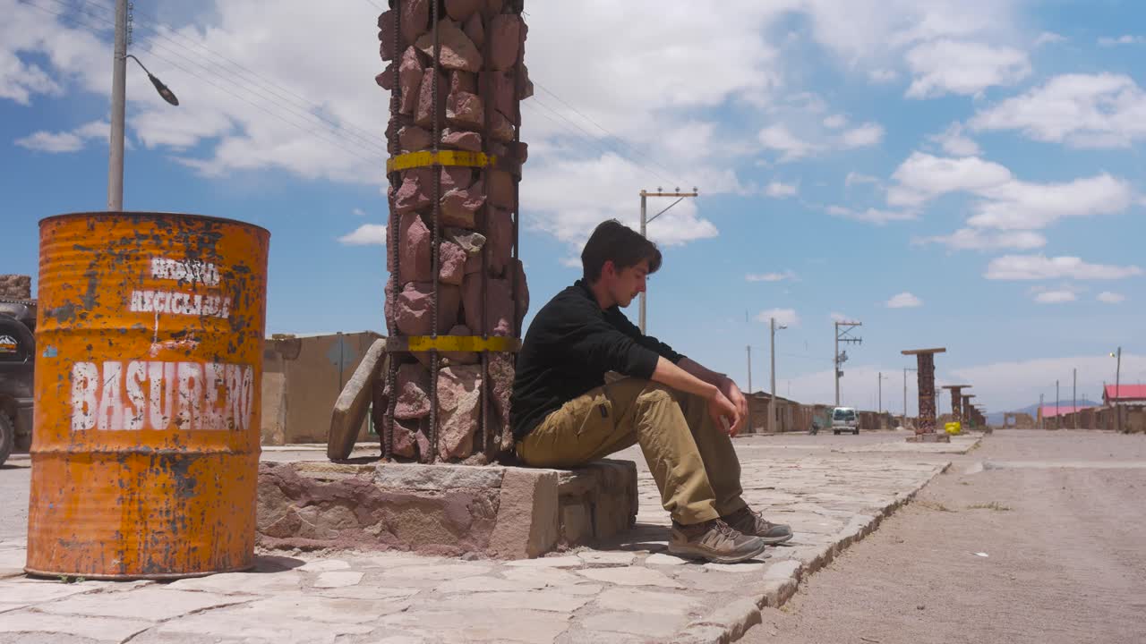 hombre solitario sentado junto a una columna de roca al lado de un tambor de basura en una calle vacía en bolivia