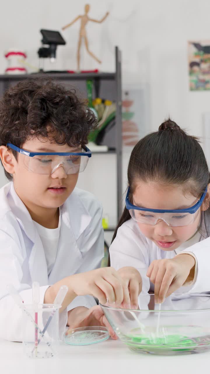 Dua siswa muda Asia mengenakan kacamata pengaman saat melakukan eksperimen sains reaksi kimia yang penuh warna.