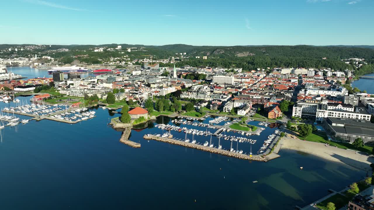 Overlooking a Kristiansand in Norway, this view showcases a marina with boats, surrounded by modern buildings and lush green hills under a clear blue sky, capturing a serene atmosphere