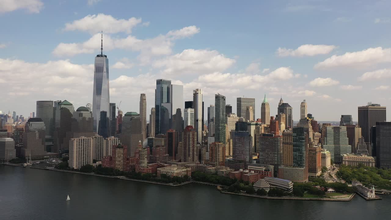 A bird's eye view over Upper Bay in New York with a single white sailboat in view. The drone hovers in place and slowly pan left to show all of lower Manhattan. It is a sunny day