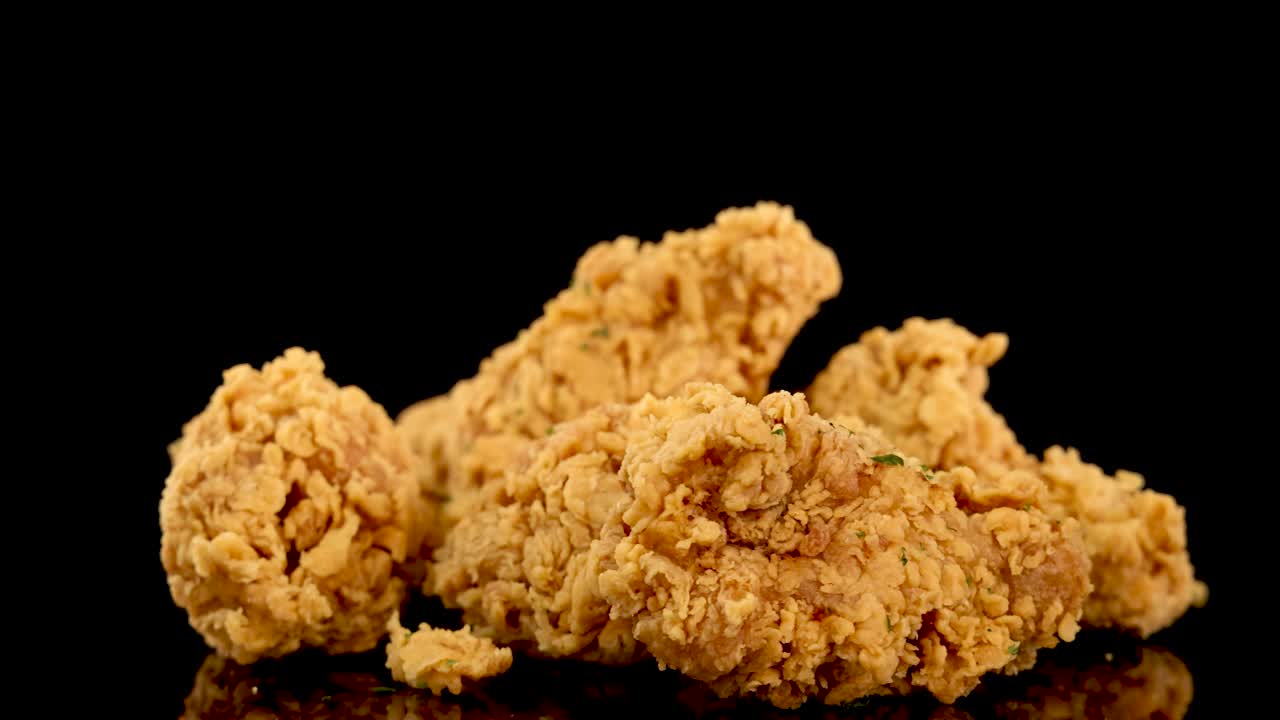 Golden, crumbed chicken breasts and thighs rotate slowly on a reflective surface with dramatic studio lighting and a deep black background