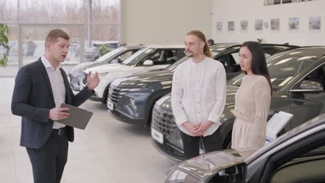 young car salesman showing to young couple new automobile at dealership salon.