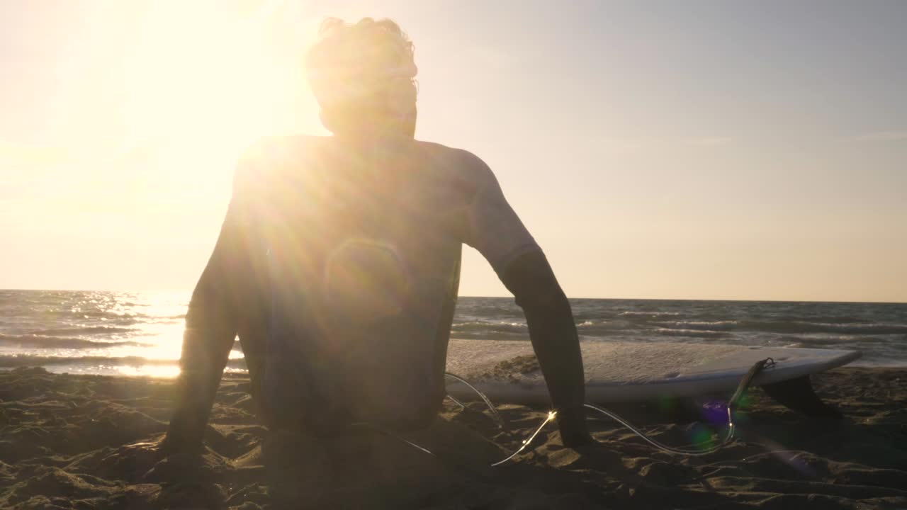 Ung mandlig surfer i våddragt sidder med surfbræt liggende på sandet på stranden, lytter til musik, danser og kigger på havet ved solnedgang.