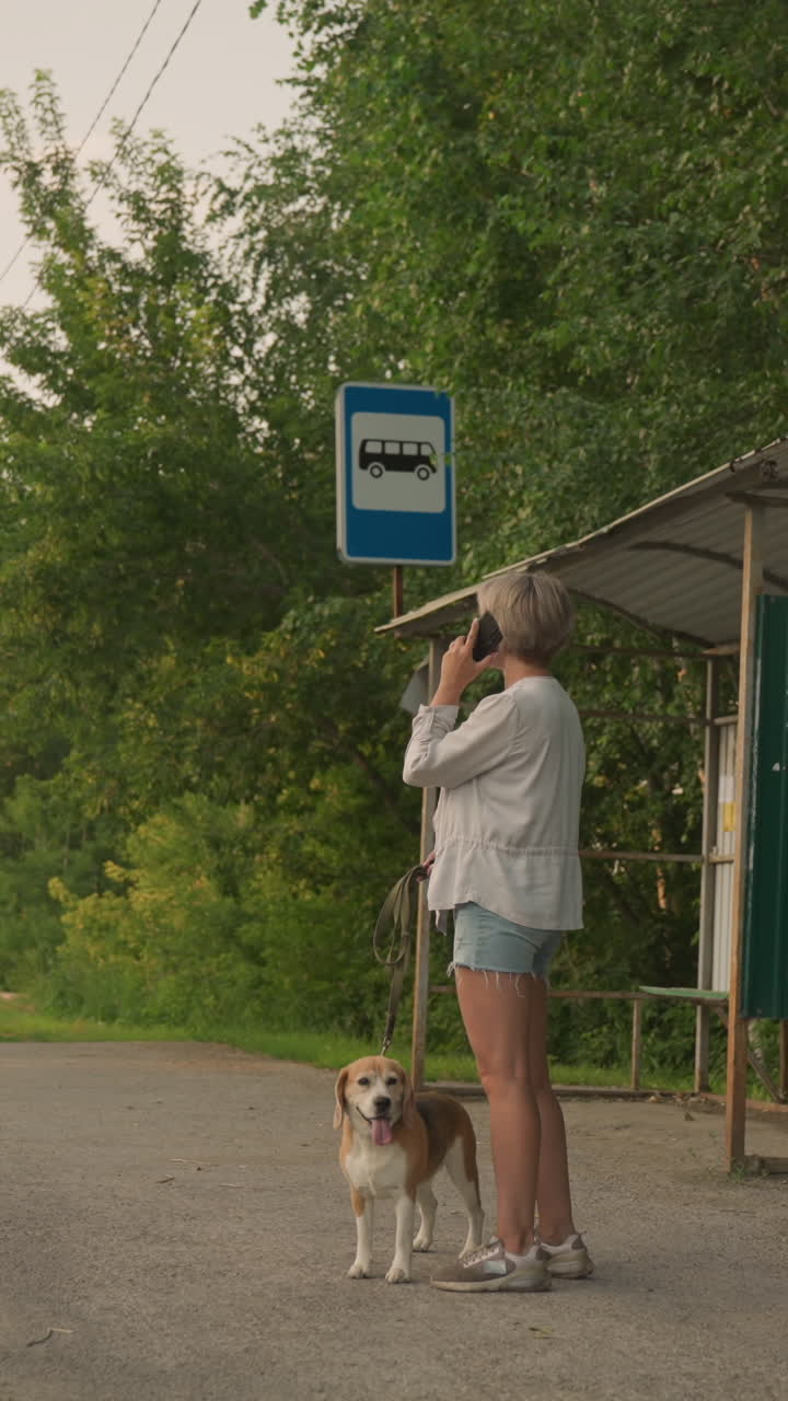 el dueño del perro de pie en la parada de autobús en una zona rural sosteniendo al perro en la correa mientras habla por teléfono, el fondo presenta árboles verdes, un refugio de parada de autobuses y una carretera con coches en el tráfico