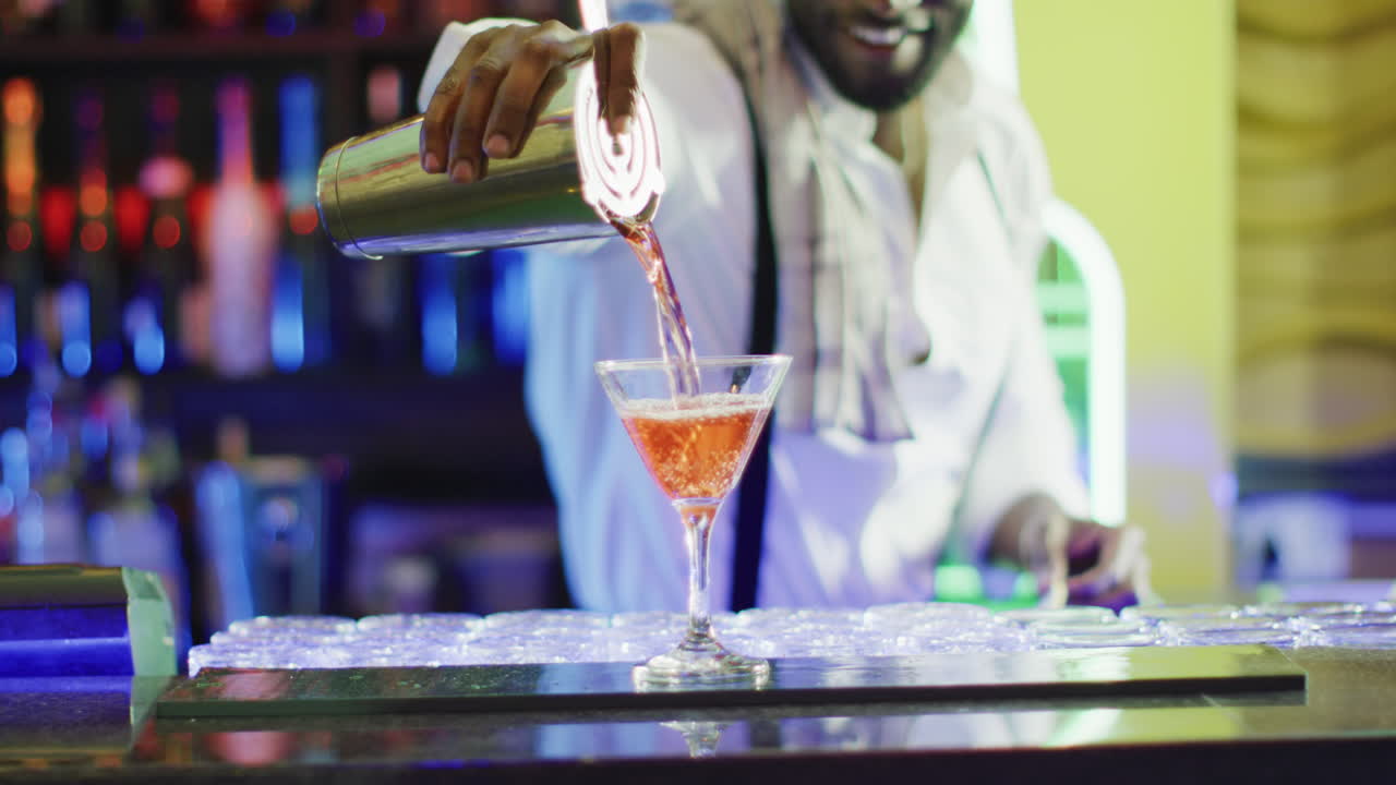 Video of midsection of smiling african american barman preparing pink cocktail at the bar