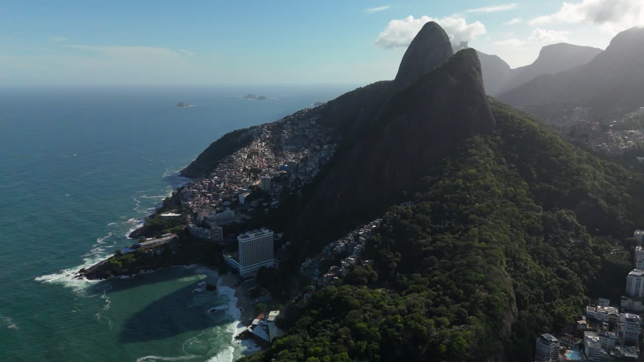 Un dron se aleja mientras asciende por la ladera de Vidigal, revelando las montañas Dos Hermanos y la costa atlántica en Río de Janeiro.