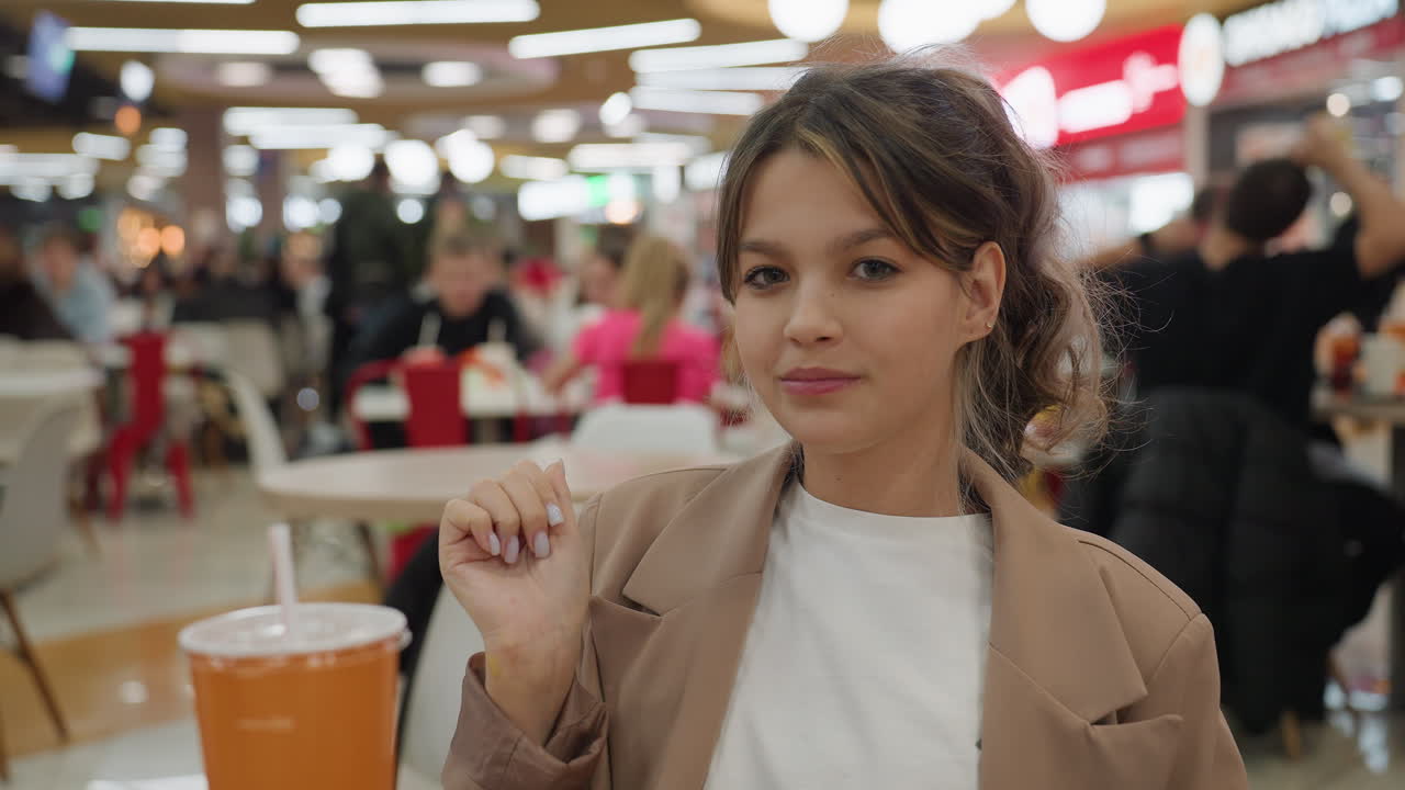 쇼핑몰 푸드코트에서 감자튀김을 다 먹고 만족스러운 표정을 짓고 있는 젊은 백인 여성, 쟁반에는 주황색 컵과 포장지가 놓여 있고 편안한 블로거 분위기, 붐비는 쇼핑객들과 머리 위 조명이 따뜻한 분위기를 연출해요