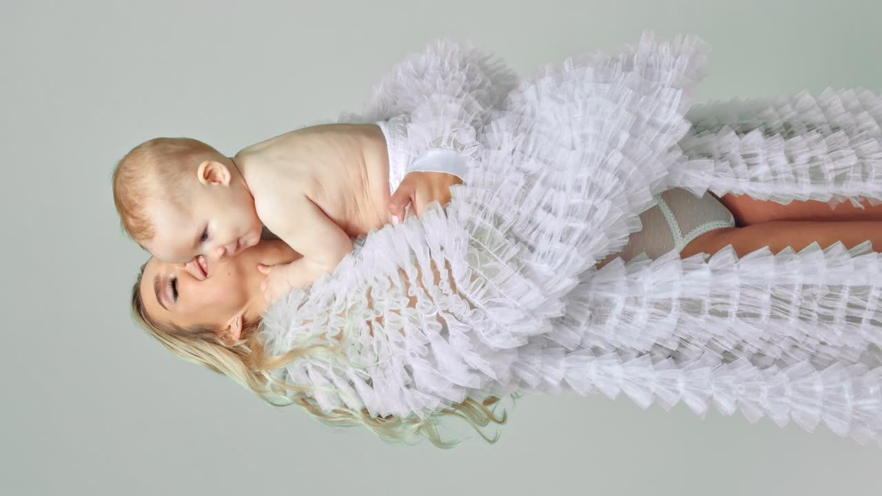 Happy young blonde woman holding a baby boy wearing a diaper. Tiny kid holding his mom by the face and kissing.