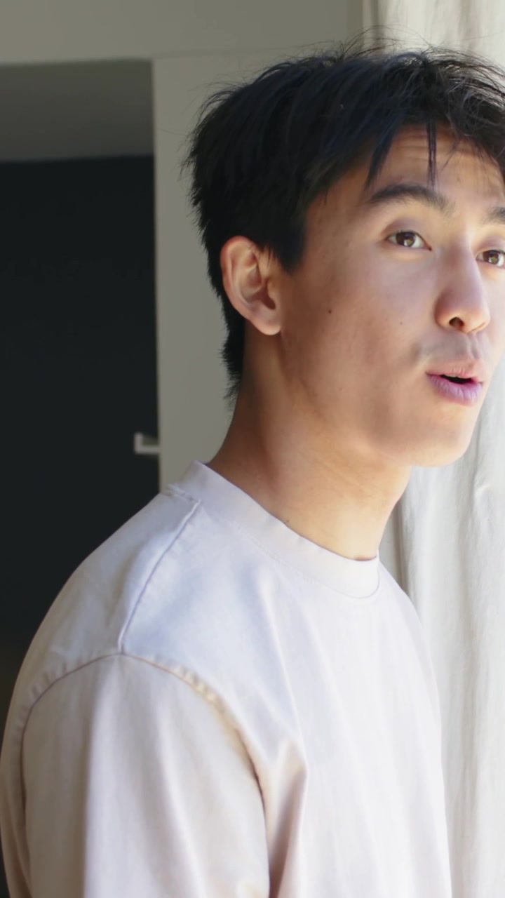 Video verticale: Uomo asiatico che si gira, traccia il movimento all'esterno accanto a una finestra, indossa una felpa beige.