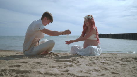 Pregnant-couple-relaxing-on-beach.-Happy-couple-pouring-sand-in-hands-on-beach