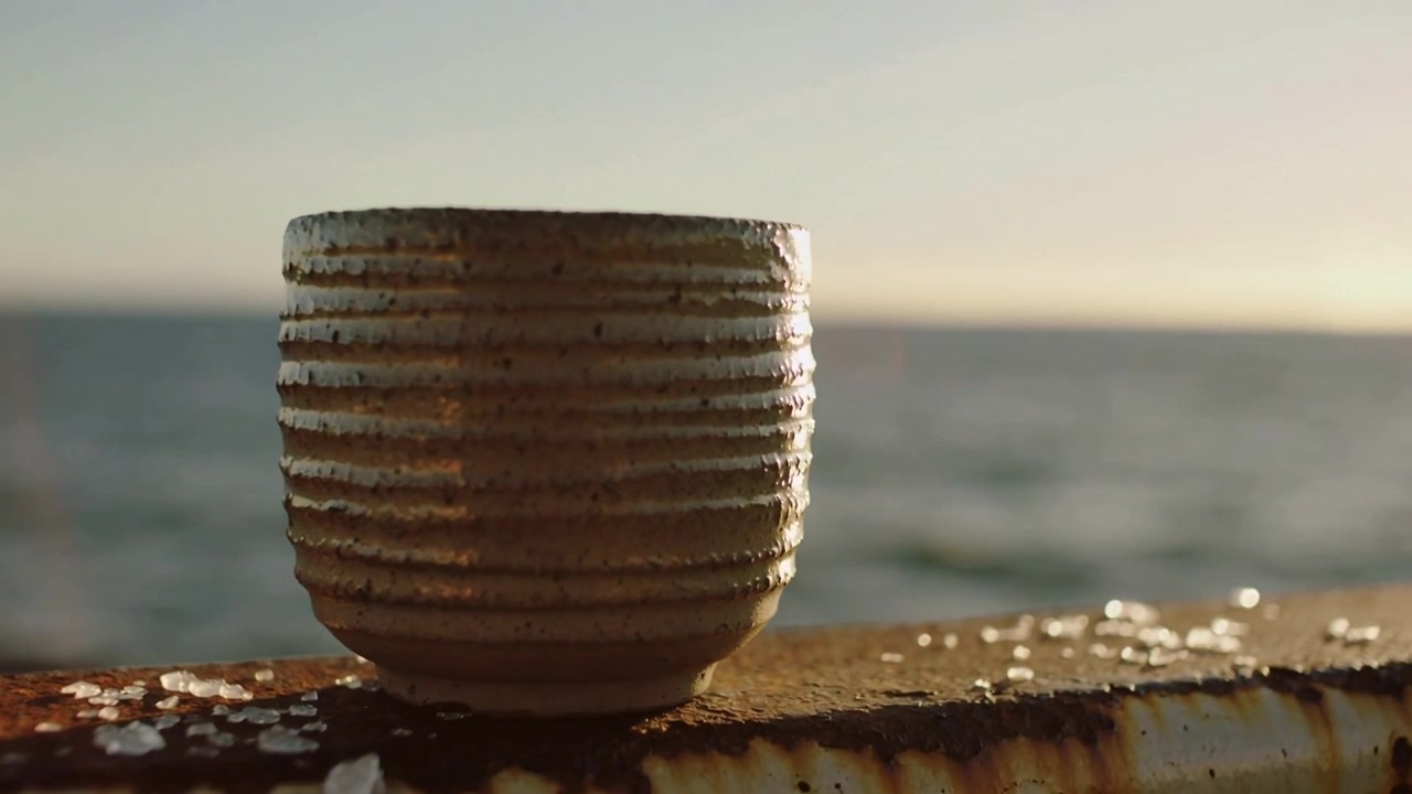 Glistening ribbed ceramic cup catching warm light on rusted pier railing with sea salt, sun moving