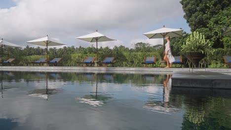 fancy blond woman in swimwear cover up dress walking along pool at bali resort, reflection on water