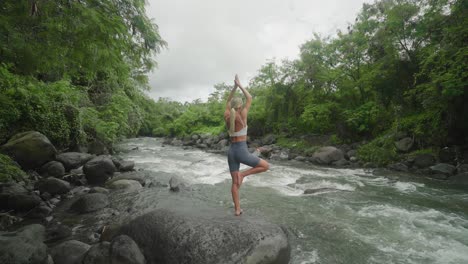 slender female practicing tree pose outdoor next to jungle river, vrksasana