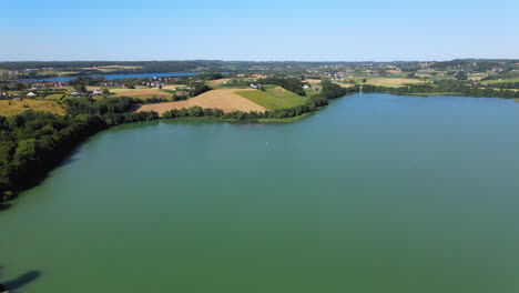 Vuelo-Aéreo-Del-Lago-Escénico-Y-Campos-Agrícolas-En-El-Fondo-Durante-El-Día-Soleado,-Polonia