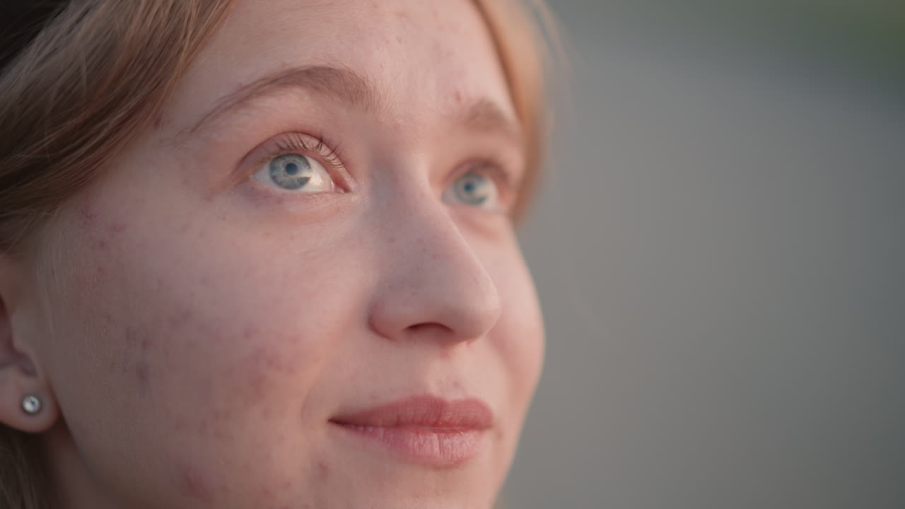 Donna caucasica sorridente che guarda il cielo, incarnato lentigginoso con orecchini che catturano una luce soffusa, primo piano spontaneo durante un festival all'aperto, bokeh ambientale e luci distanti, occhi luminosi e delicati
