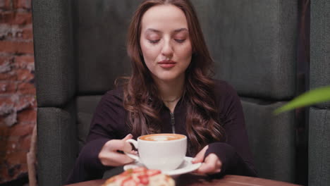 young woman in dark outfit gently lifts latte toward face with eyes closed, enjoying aroma beside plate of waffles topped with strawberries, in cozy setting with wooden table and soft lighting