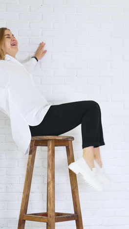 long-haired lady in white shirt sits on a high stool waving her legs. white modern shoes presentation at the backdrop of white brick wall. vertical video
