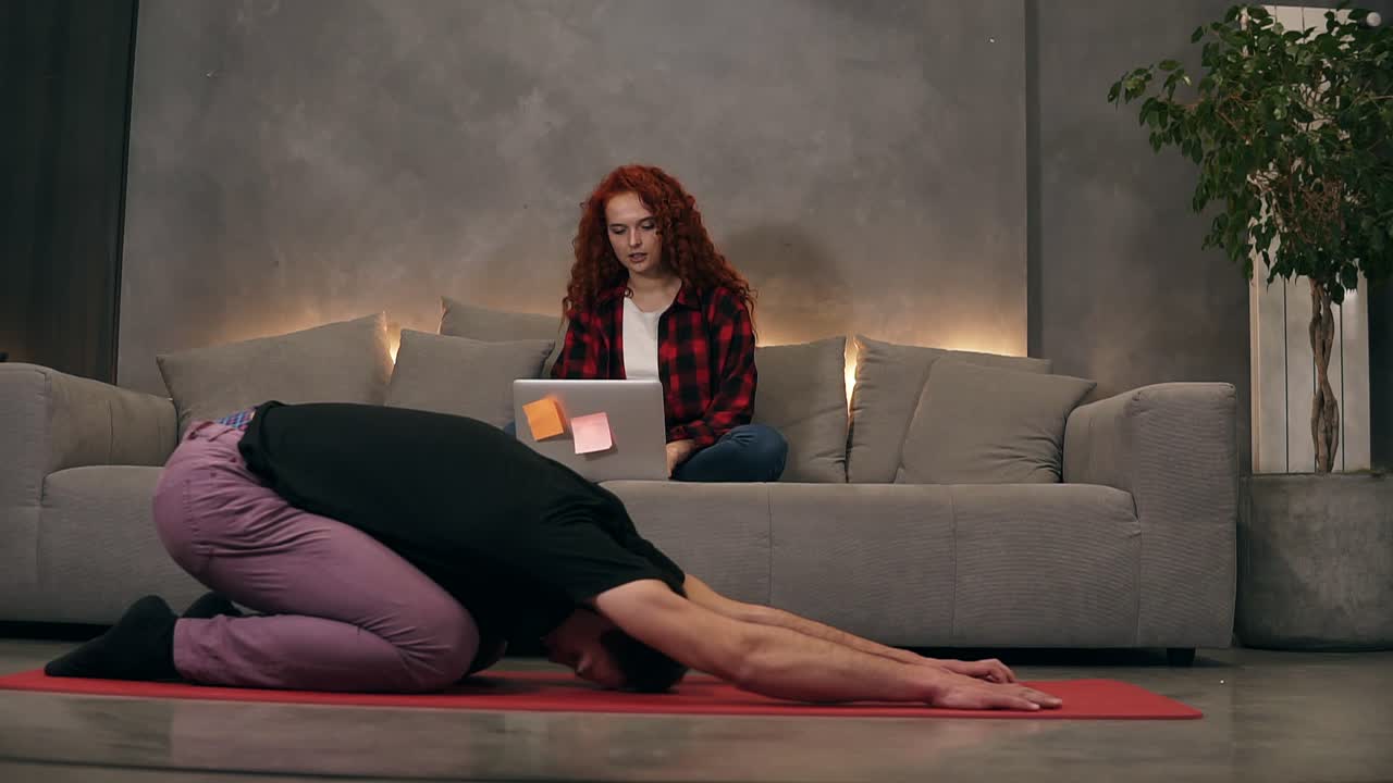 Young caucasian couple at relaxing home living room, curly readheaded firl using modern wireless laptop on a grey couch while her boyfriend is doing yoga, stretching on a red mat. Front view. Loft designed living room